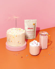 Blume Superfood Birthday Cake Latte Blend with a cake, hot and iced latte on a orange countertop. 